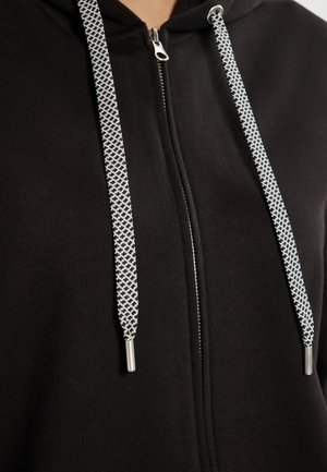 Haut de sweatshirt zippé noir avec une texture lisse, cordons blancs avec un motif en diamant, et accents en métal argenté aux extrémités.