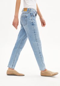 Vaaleansiniset farkut suoralla lahkeella, viidellä taskulla ja ruskea nahkainen merkki vyötäröllä, yhdistettynä ruskeisiin slip-on-kenkiin.