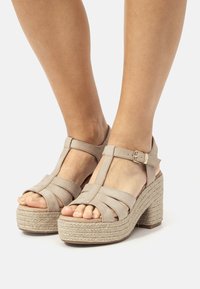 Beige High Heels Sandalen mit einem T-Riemen-Design, aus weichem Wildleder, mit einer gewebten Espadrille-Plattform und einem mit einer Schnalle verstellbaren Knöchelriemen.