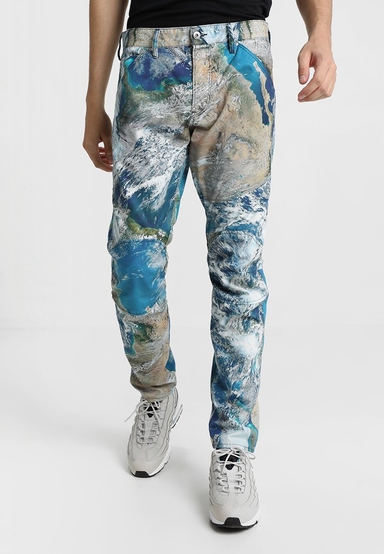 Man die een spijkerbroek draagt met een realistische print van een wereldkaart, gecombineerd met witte sneakers en een zwart shirt, staande op een effen witte achtergrond.