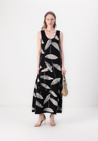 Maxiklänning i svart med vitt lövmönster. Har rund halsringning, är ärmlös och har en lös passform. Modellen håller en vävd väska.