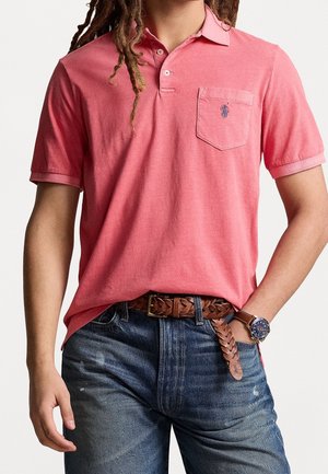 Mand iført et koralrosa polo shirt med brystlomme, flettet brun læderbælte, blå jeans og brunt ur med blå skive, hånd i lommen.