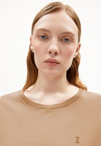 Braunes Baumwoll-T-Shirt mit rundem Ausschnitt und kurzen Ärmeln. Verfügt über ein kleines, tonal gehaltenes Logo auf der Brust. Glatte Textur.