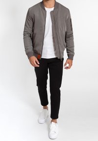 Man wearing gray bomber jacket over white t-shirt, black pants, and white sneakers, walking against a plain white background.