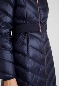 Abrigo acolchado de color azul marino con un patrón en forma de chevrón, que presenta un cinturón ancho y texturizado y una cremallera de contraste. Fabricado con un tejido brillante y de textura suave.