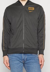 Svart zip-up jacka med orange detaljer, Nike-logotyp på vänster bröst och sidofickor. Tillverkad av ett mjukt, flexibelt tyg.