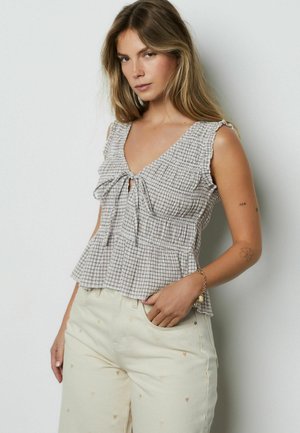 Grauer und weiß kariertes Peplum-Oberteil mit V-Ausschnitt, Rüschenträgern und vorderer Schleifendetail, kombiniert mit cremefarbenen Hosen mit kleinen Herzmustern.