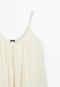 Witte mouwloze top met dunne bandjes, geplooide halslijn en loshangende strikdetails aan de voorkant.