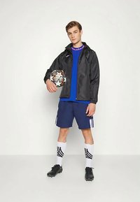 Black lightweight jacket over a blue shirt, paired with navy shorts, white socks featuring black stripes, and black soccer cleats. Holding a soccer ball.
