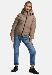 Beige pufferjack met diagonaal gestikt patroon, voorrits, trekkoordkapuchon en zijzakken. Draagt met lichtblauwe spijkerbroek en zwarte sneakers.