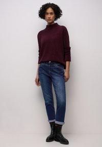 Suéter de punto color granate con cuello alto, combinado con vaqueros azules con los puños doblados y botines negros. Texturas suaves, ajuste casual.