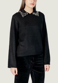 Suéter de punto negro con mangas largas y un acabado texturizado. El cuello cuenta con adornos de cuentas blancas como acento. Combinado con pantalones de terciopelo negro.