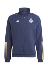 Veste de sport bleu marine zippée avec le logo du Real Madrid, le logo Adidas, et des bandes dorées, blanches et bleu marine sur les manches.