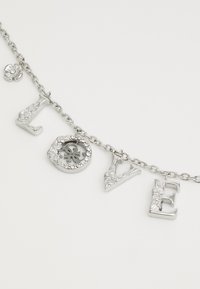 Pulsera de cadena plateada con letras entrelazadas que forman la palabra "AMOR." Presenta letras texturizadas y un charm circular adornado con rhinestones.