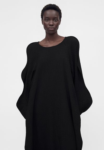 Γυναίκα με κοντά σγουρά μαλλιά φορώντας μια φαρδιά μαύρη υφασμάτινη φόρεμα με φουσκωτά μανίκια μπροστά σε ένα απλό φωτεινό φόντο.