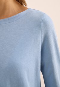 Haut bleu clair à manches longues avec un large décolleté, une texture douce et un motif de tissu subtil et uniforme. Met en valeur des bords bruts au niveau du décolleté.