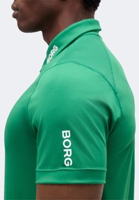 Άνδρας με πράσινο πόλο πουκάμισο με το λογότυπο "BORG" σε λευκό στο γιακά και το μανίκι, εμφανίζεται από το πλάι με ουδέτερο φόντο.