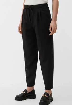 Femme portant un pantalon noir fuselé avec une taille à cordon de serrage et des mocassins noirs avec un détail de chaîne dorée, debout devant un fond blanc.