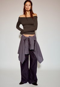 Długi rękaw, bluzka z odkrytymi ramionami w ciemnym brązie, zestawiona z luźnymi, ciemnymi dżinsami oraz koszulą w kratę zawiązaną w pasie.