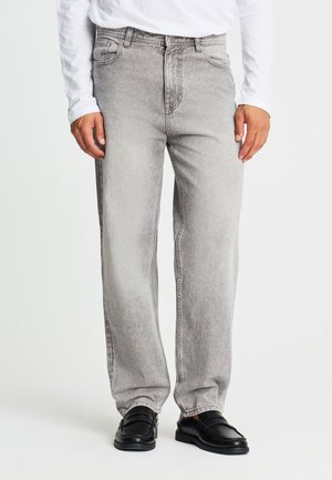 Uomo che indossa jeans grigio chiaro a gamba dritta, mocassini neri e una camicia bianca a maniche lunghe, in piedi davanti a uno sfondo semplice.