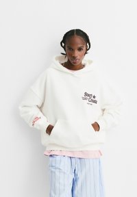 Witte fleece hoodie met een voorzak, omgeslagen mouwen en bedrukte tekst op de borst en mouw in bruin en rood. Draag de hoodie over een roze shirt.