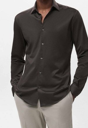 Homme portant une chemise ajustée gris foncé à boutons avec col et un pantalon beige, mains dans les poches, debout sur un fond uni.