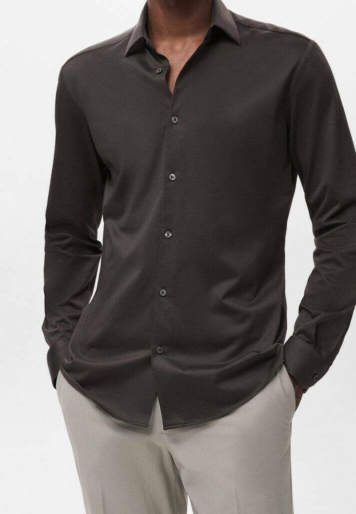 Homme portant une chemise ajustée gris foncé à boutons avec col et un pantalon beige, mains dans les poches, debout sur un fond uni.