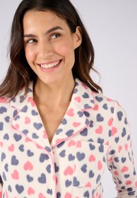 Fleece pyjama-top met een crèmekleurige achtergrond, versierd met een hartjespatroon in roze en marineblauw. Inclusief een borstzak en roze bies op de kraag.