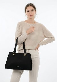 Femme portant un pull beige et un pantalon, tenant un grand sac à main noir avec double anse sur son bras gauche, debout devant un fond blanc.