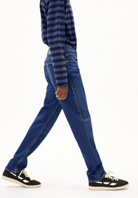 Denim jeans in een middelblauwe wassing met een rechte pijp, gedragen met een donker gestreepte lange mouwen shirt en zwarte sneakers.