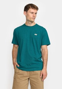 Camiseta de algodón color teal con cuello redondo y mangas cortas. Presenta un pequeño parche con logo en el pecho. Corte casual. Combinada con pantalones cortos de carga beige.