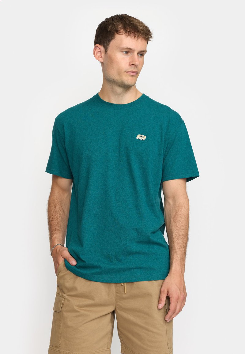 Camiseta de algodón color teal con cuello redondo y mangas cortas. Presenta un pequeño parche con logo en el pecho. Corte casual. Combinada con pantalones cortos de carga beige.