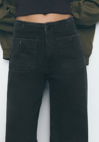 Schwarze, hoch taillierte Jeans mit geradem Bein, ausgestattet mit zwei großen Vorderstaschen und Metallknopfverschlüssen. Glatte Denim-Oberfläche.