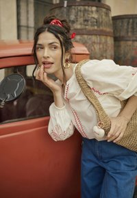 Donna con i capelli scuri legati da un foulard rosso, che indossa una camicetta bianca ricamata e jeans, appoggiata a un'auto rossa d'epoca vicino a delle botti di legno.