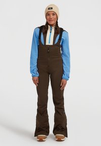 Salopette da sci marroni con bretelle regolabili, abbinata a un fleece blu e una giacca zip crema. Il modello indossa un berretto beige e stivali da neve.