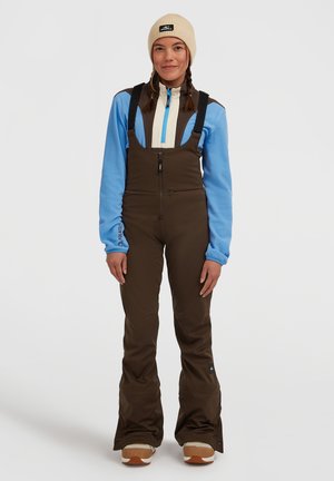 Bruine skibroek met verstelbare banden, gecombineerd met een blauwe fleece en een crèmekleurige ritsjack. Het model draagt een beige beanie en sneeuwschoenen.