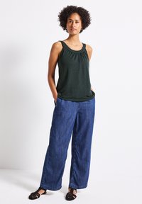 Haut sans manches vert foncé avec un col froncé, associé à un pantalon en denim bleu à jambe large. Le mannequin se tient avec les mains dans les poches devant un fond blanc.