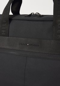 Sac noir Tommy Hilfiger avec fermeture à double zip, poignées de marque, et une bande horizontale en tissu avec le logo Tommy Hilfiger.
