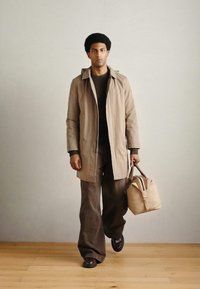 Bézs vízálló kabát sötét pulóver fölött, barna bőszárú nadrággal párosítva. Világos bézs táska, csomózott részlettel. Barna cipő.