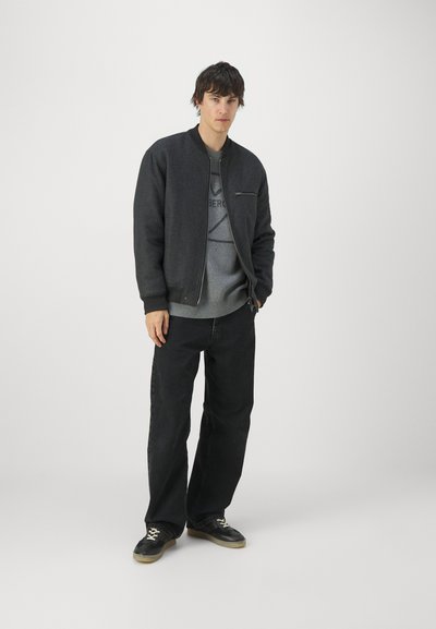 Chaqueta bomber de color gris oscuro con cuello redondo, cierre con cremallera y tejido texturizado, superpuesta a un suéter gris claro y jeans negros.