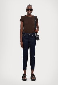 Καφέ υφασμάτινη μπλούζα κοντομάνικη, μπλε τζιν παντελόνια μέχρι τον αστράγαλο, και μαύρα σανδάλια χωρίς φάσα. Τα αξεσουάρ περιλαμβάνουν μια μικρή μαύρη τσάντα χιαστί και γυαλιά ηλίου.