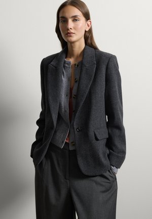 Mujer con cabello castaño liso lleva una chaqueta gris oscuro sobre un cárdigan gris abotonado y pantalones oscuros sueltos, de pie frente a una pared lisa y clara.