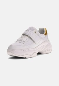 Gruby biały sneaker z grubą podeszwą, sznurków z przodu, szerokim paskiem na rzep oraz złotym akcentem na kołnierzu pięty, ustawiony pod kątem w lewo.