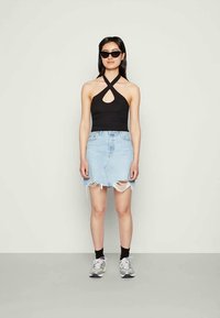 Mulher vestindo um top preto com decote halter, saia de ganga desfiada de um azul claro, meias pretas, ténis cinzentos e óculos de sol pretos com formato de gato em pé contra um fundo simples.