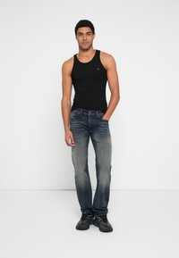 Μαύρο βαμβακερό αμάνικο μπλουζάκι, μπλε τζιν παντελόνι με ξεθωριασμένη υφή και μαύρα αθλητικά παπούτσια. Το άτομο στέκεται όρθιο σε λευκό φόντο.