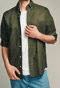 Camicia button-up verde oliva in cotone, con logo sul petto, polsini abbottonati e vestibilità relaxed sopra una t-shirt bianca e jeans in denim.