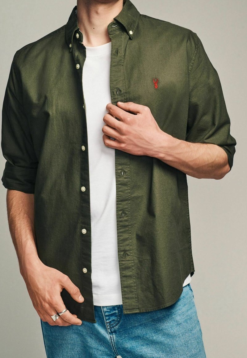 Camicia button-up verde oliva in cotone, con logo sul petto, polsini abbottonati e vestibilità relaxed sopra una t-shirt bianca e jeans in denim.