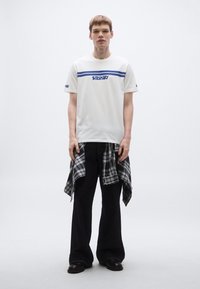 Bela majica s modrimi črtami in logotipom, kombinirana s črnimi jeans hlačami. Črno-bela karo srajca je povezana okoli pasu. Črni čevlji.