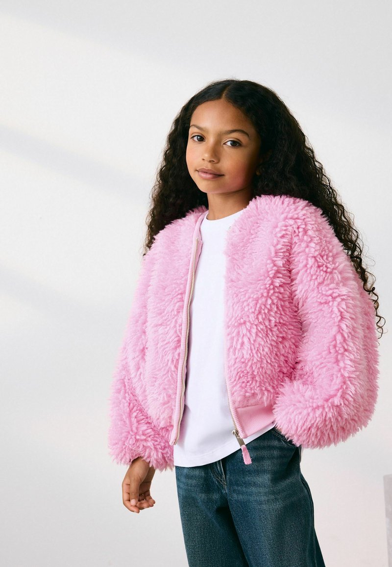 Jeune fille aux longs cheveux bouclés portant une veste rose duveteuse, une chemise blanche et un jean bleu, debout devant un fond clair uni.