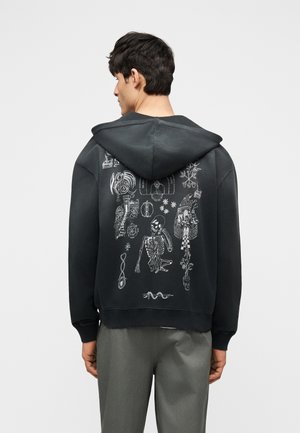Młody mężczyzna w czarnej bluzie z kapturem z białymi abstrakcyjnymi szkicami i szkicami szkieletu na plecach, ubrany w szare spodnie, stojący na białym tle.